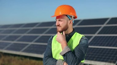 Mühendis ayakta kameraya bakıyor. O, elini sakalına dayıyor. Arka planda güneş panelleri var. 4K