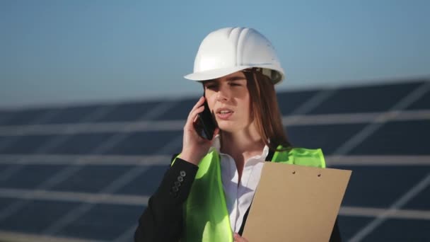 En kvinnlig ingenjör pratar i telefon. Hon håller dokument i sina händer. Hon står nära solpanelerna. 4K — Stockvideo