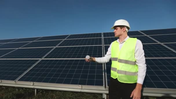 O engenheiro está ao lado dos painéis solares. Ele está pegando a lâmpada e olhando para ela. 4K — Vídeo de Stock