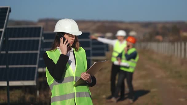 Жінка-інженер розмовляє по телефону. Двоє інженерів говорять на задньому плані. Вона стоїть біля сонячних панелей. 4-кілометровий — стокове відео