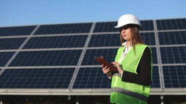 Kadın mühendis objeyi inceliyor ve veriyi tablete giriyor. Güneş panellerinin yanında duruyor. 4K