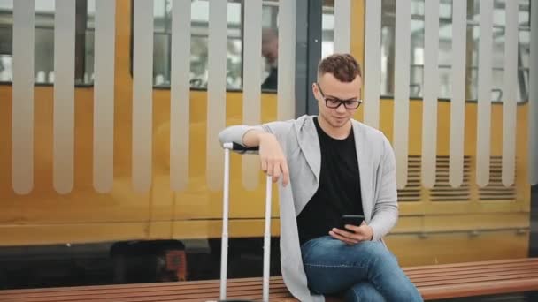 Un hombre está sentado en una parada de transporte público. Él está enviando mensajes de texto en su teléfono inteligente y sosteniendo una maleta. Tranvía en el fondo. 4K — Vídeo de stock