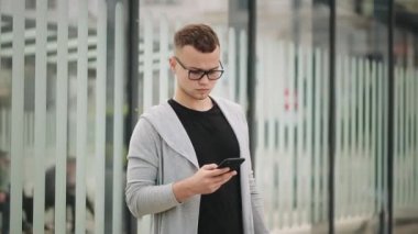 Bir adam toplu taşıma durağında duruyor ve etrafa bakıyor. O, akıllı telefonuyla mesaj atıyor. 4K