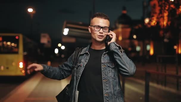 Un hombre está hablando emocionalmente por teléfono. Está parado en una parada de transporte público. Ciudad Nocturna al fondo. 4K — Vídeos de Stock