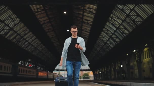Mężczyzna idzie wzdłuż peronu stacji. Trzyma walizkę. Patrzy na smartfona i sprawdza wiadomości. 4K — Wideo stockowe