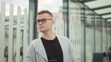 Bir adam toplu taşıma durağında duruyor. Etrafa bakıyor. O gözlük takıyor. 4K