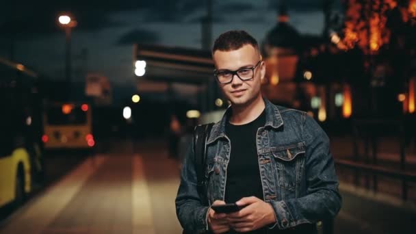 Ein Mann schreibt eine SMS auf seinem Smartphone. Er steht an einer Haltestelle des öffentlichen Nahverkehrs. Night City im Hintergrund. 4K — Stockvideo
