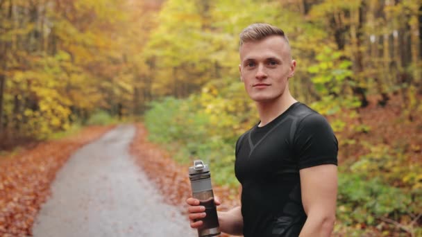 El atleta está de pie en un camino forestal y mirando a la cámara. Está sosteniendo una botella de agua y levantando el dedo. Ejecutando entrenamiento. 4K — Vídeos de Stock