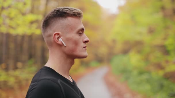 Sportowiec stoi na leśnej drodze. Słucha muzyki ze słuchawkami na uszach i odwraca głowę. Patrzy w kamerę. 4K — Wideo stockowe