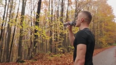 Sporcu bir orman yolunda duruyor ve bir şişeden su içiyor. Sonbahar ormanında koşu antrenmanı. Kamera onun etrafında dairesel bir hareket yapıyor. 4K
