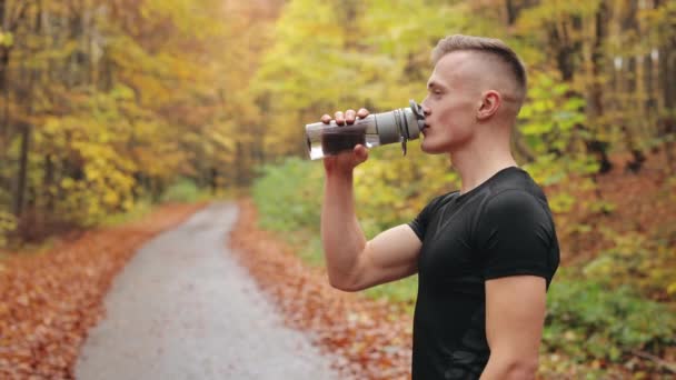 A sportoló egy erdei úton áll, és egy üvegből issza a vizet. Elfordítja a fejét és a kamerába néz. Edzést tartok. 4K — Stock videók