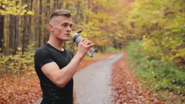 Sportowiec stoi na leśnej drodze i pije wodę z butelki. Trening biegowy w jesiennym lesie. 4K — Wideo stockowe