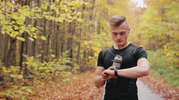De atleet staat op een bosweg en controleert de trainingsgegevens op het sporthorloge. Hardlooptraining in het herfstbos. 4K — Stockvideo