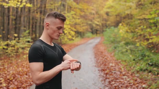 Sportowiec stoi na leśnej drodze i sprawdza dane treningowe na zegarku sportowym. Podnosi głowę i patrzy w kamerę. Trening biegowy w jesiennym lesie. 4K — Wideo stockowe