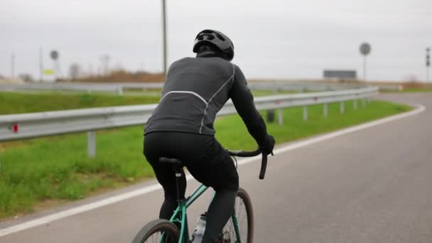 Спортсмен тренируется на велосипеде. Он едет по шоссе в холодное время года. Он делает поворот. Стрелял сзади. 4K — стоковое видео