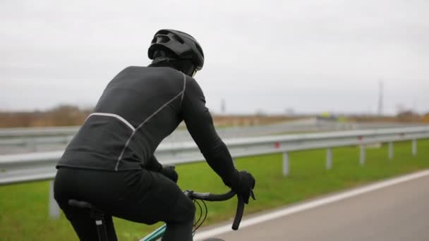 Спортсмен тренируется на велосипеде. Он едет по шоссе в холодное время года. Он одевается в тёплую одежду. Стрелял сзади. 4K — стоковое видео