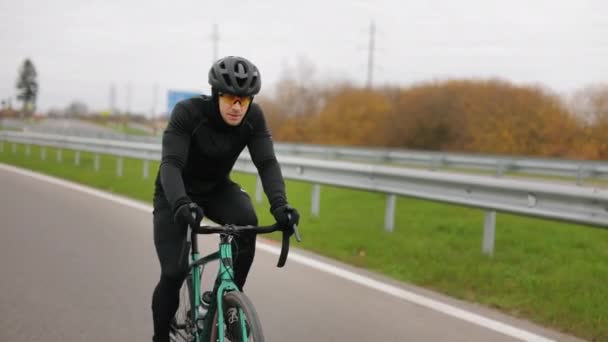 The athlete is training on a bicycle. He is driving on the highway in the cold season. He is dressing in warm gear. 4K — Stock Video