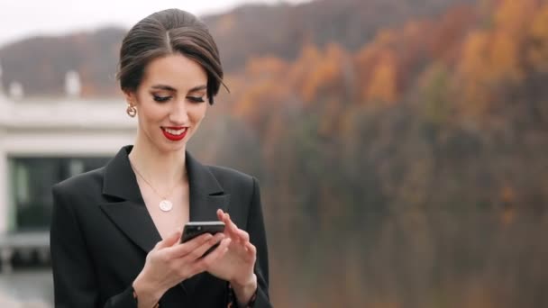 Femeia este în picioare și mesaje text pe smartphone-ul ei. Zâmbeşte. E îmbrăcată elegant. 4K — Videoclip de stoc