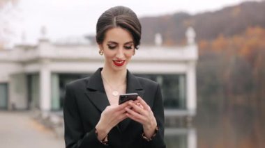 Kadın ayakta duruyor ve akıllı telefonuyla mesaj atıyor. O gülümsüyor. O şık giyinmiş. O, gölün yanında duruyor. 4K