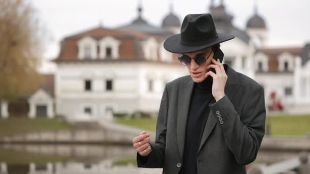 Omul se duce şi vorbeşte la telefon. E îmbrăcat elegant. El poartă o pălărie. Lacul în fundal. 4K — Videoclip de stoc