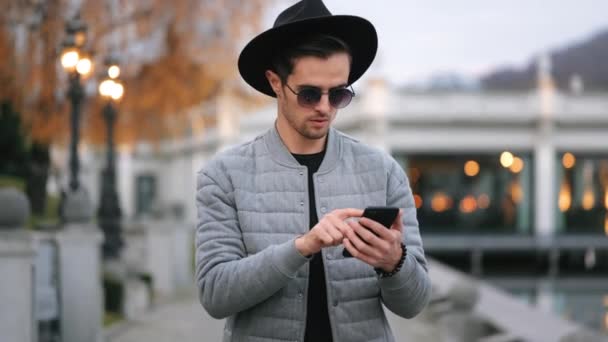 Ein Mann geht am Kai entlang und schreibt SMS auf seinem Smartphone. Er ist stilvoll gekleidet. Er trägt Hut und Sonnenbrille. Der schöne Kai im Hintergrund. 4K — Stockvideo
