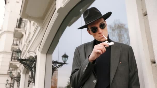 Der Mann steht und zündet sich mit einem Feuerzeug eine Zigarette an. Er lehnt an der Wand. Er ist stilvoll gekleidet. Er trägt eine Sonnenbrille und einen schwarzen Hut. 4K — Stockvideo
