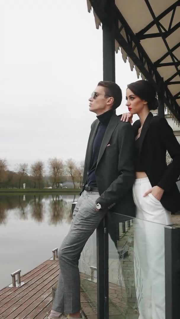 Чоловік і жінка стоять і дивляться на озеро. Вони повертаються і дивляться на камеру. Вони стильно одягнені. Вертикальне відео . — стокове відео