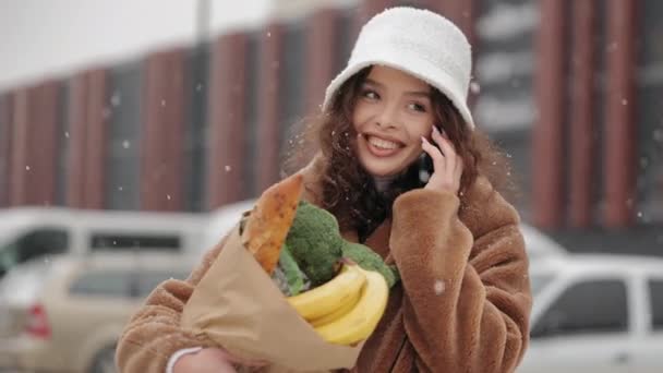 Een vrouw staat in de buurt van een supermarkt en praat op een smartphone. Ze glimlacht en houdt een boodschappentas vast. Zware sneeuwval. Portret schieten. 4K — Stockvideo