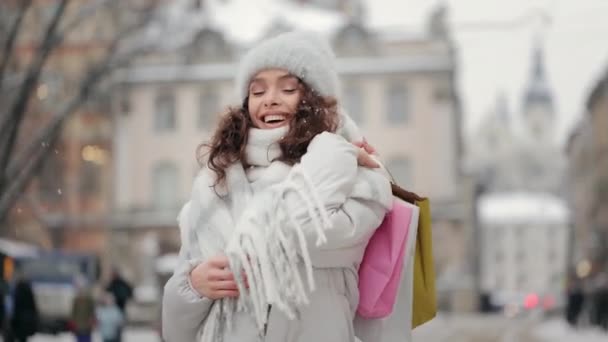 Uma mulher está de pé no centro da cidade e sorrindo. Ela está olhando para a câmera e segurando sacos de compras em seu ombro. A neve está a cair. 4K — Vídeo de Stock