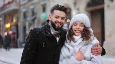 Bir adam ve bir kadın bir kar yağışı altında şehrin merkezinde duruyorlar. Sarıldılar ve kameraya bakıyorlar. Portre çekimi. 4K