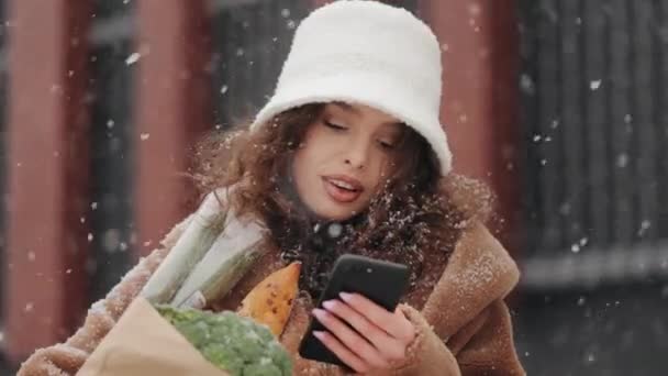 Une femme se tient près d'un supermarché et envoie des SMS sur un smartphone. Elle lève la tête et regarde la caméra. Elle sourit et tient un sac d'épicerie. La neige tombe. Portrait — Video