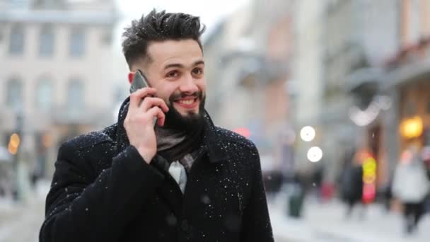 Un homme se tient sur la place centrale dans une chute de neige. Il parle sur son smartphone et sourit. Tournage de portraits. 4K — Video