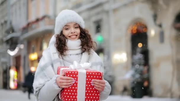 Uma mulher está em pé em uma praça coberta de neve da cidade. Ela está segurando um grande presente e olhando para a câmera. Ela está sorrindo. Ela está a gostar do tempo. 4K — Vídeo de Stock