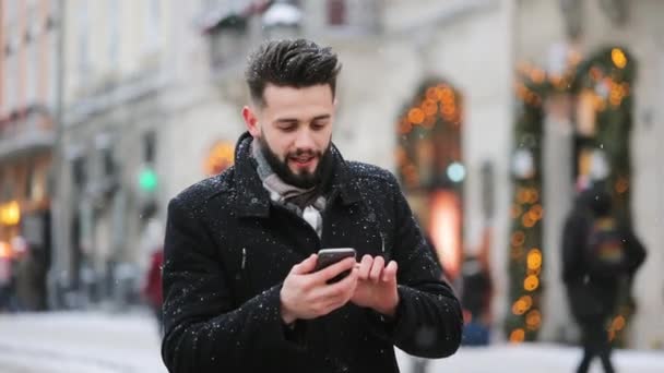 한 남자 가 중앙 광장에 눈이 내리고 있습니다. 그는 스마트폰으로 문자를 보내며 웃고 있습니다. 4K — 비디오
