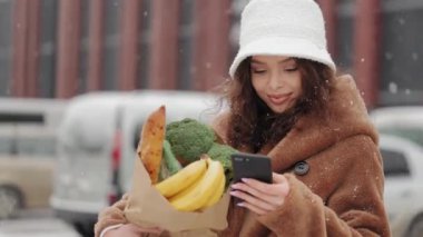 Bir kadın süpermarketin yanında duruyor ve akıllı telefondan mesaj atıyor. O gülümsüyor ve bir market poşeti tutuyor. Yoğun kar yağıyor. 4K