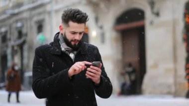 Merkez meydanda kar yağışı altında bir adam duruyor. Akıllı telefonuyla mesaj atıyor ve gülümsüyor. 4K