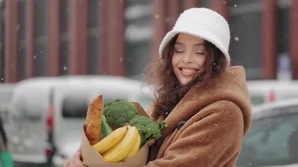 Жінка стоїть біля супермаркету і дивиться на камеру. Вона посміхається і тримає продуктовий пакет. Сильний сніг падає. Машина їде на задньому плані. 4-кілометровий — стокове відео