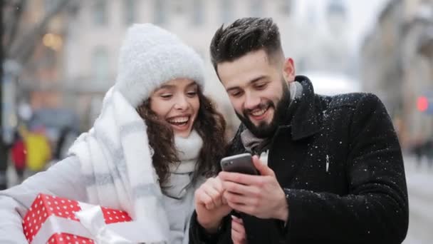 Egy férfi és egy nő áll a város közepén egy hóesésben. Egy nő ajándékot tart a kezében. Az okostelefonjukat nézik és nevetnek. Portré fotózás. 4K — Stock videók