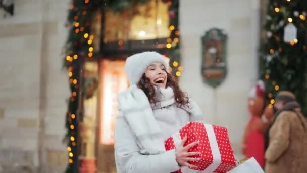 Eine Frau steht während eines Schneefalls in der Innenstadt. In den Geschäften liegen ein großes Geschenk und Einkaufstüten. Sie lächelt und springt. Porträtschießen. 4K — Stockvideo