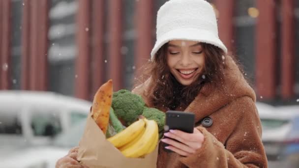 Жінка стоїть біля супермаркету і надсилає смс на смартфон. Вона піднімає голову і дивиться на камеру. Вона посміхається і тримає продуктовий пакет. Сильний сніг падає. 4-кілометровий — стокове відео