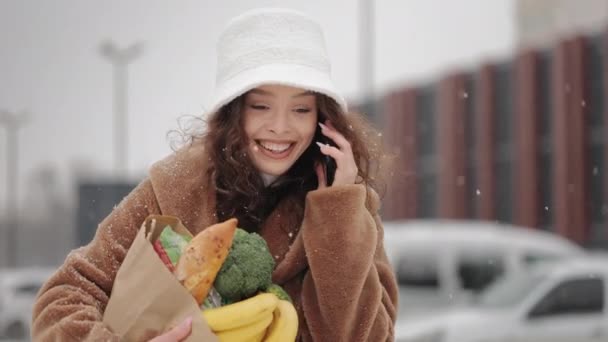 Жінка йде біля супермаркету і розмовляє по смартфону. Вона посміхається і тримає продуктовий пакет. Сильний сніг падає. 4-кілометровий — стокове відео