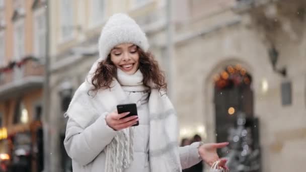 Uma mulher está de pé no centro da cidade e mensagens de texto em um smartphone. Ela está sorrindo. Ela está olhando para a câmera e segurando sacos de compras em seu ombro. A neve está a cair. 4K — Vídeo de Stock