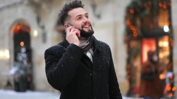 在一场大雪中，一个人站在中央广场上。他在用智能手机说话，面带微笑。4K — 图库视频影像