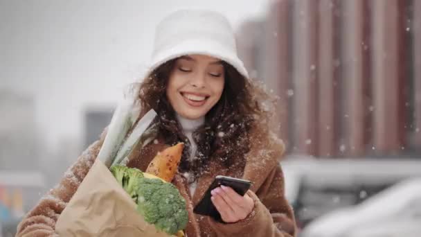 Kobieta przechodzi przez parking w pobliżu supermarketu i pisze smsy na smartfonie. Podnosi głowę i patrzy w kamerę. Uśmiecha się i trzyma torbę z zakupami. Duże opady śniegu — Wideo stockowe