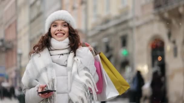 Kobieta idzie do centrum miasta i pisze smsy na smartfonie. Uśmiecha się i trzyma torby na zakupy. Patrzy w przyszłość z satysfakcją. Śnieg pada. 4K — Wideo stockowe