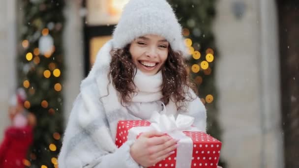 Egy nő áll a város hófödte terén. Nagy ajándékot tart a kezében, és a kamerába néz. Mosolyog. Élvezi az időjárást. Portré fotózás. 4K — Stock videók