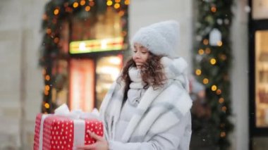 Şehrin karla kaplı bir meydanında bir kadın duruyor. Büyük bir hediye veriyor ve gülüyor. Portre çekimi. 4K