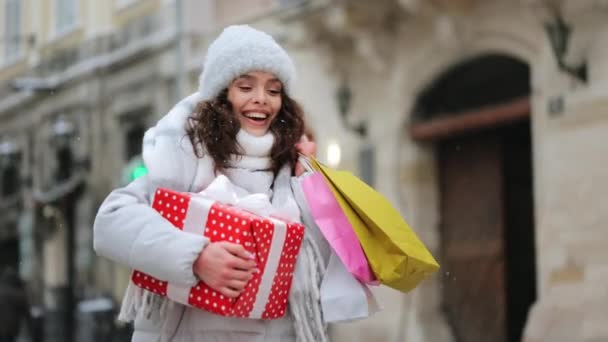 Uma mulher vai para o centro da cidade durante uma queda de neve. Ela está segurando um grande presente e sacos de compras. Ela está sorrindo. 4K — Vídeo de Stock