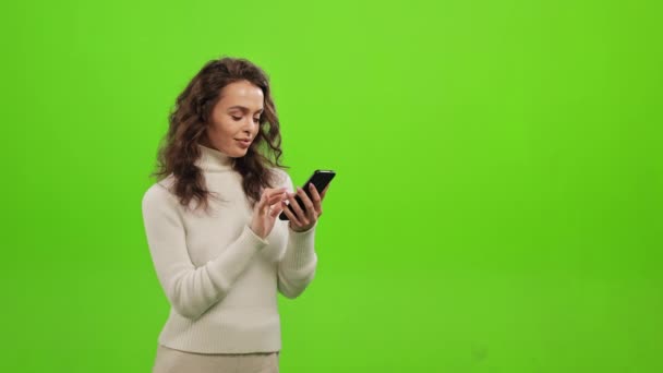 Die Frau schaut auf ihrem Smartphone durch die sozialen Netzwerke. Sie hebt den Kopf und lächelt. Sie steht auf grünem Grund. Grüner Bildschirm. 4K — Stockvideo