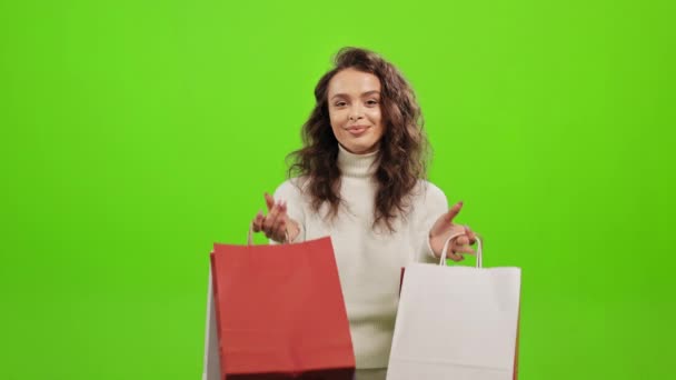 A mulher está a olhar para a câmara e a sorrir. Ela vai buscar os sacos de compras. Ela está de pé sobre um fundo verde. Tela verde. 4K — Vídeo de Stock